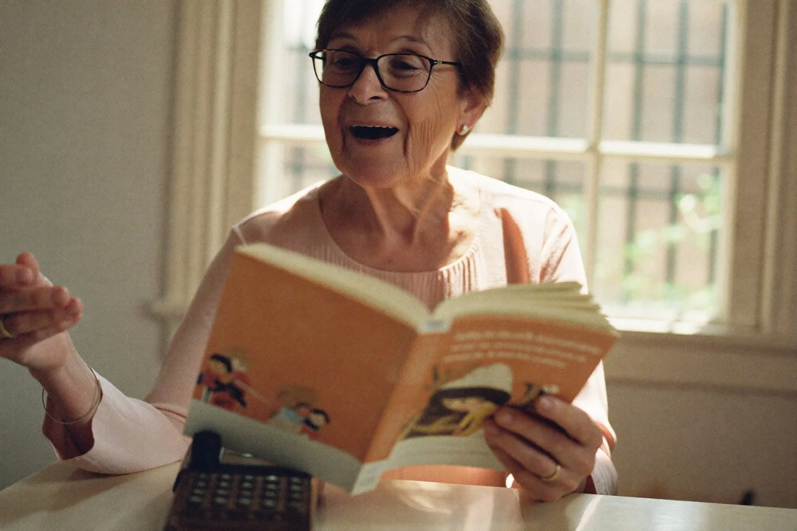 A grandmother at a sunlit kitchen table reading a picture book aloud to her phone